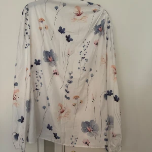 Ljus blus med blommigt mönster - Elegant och feminin blus i en mjuk, lätt tygkvalitet. Vit bas med vackra blommor i dova blå, rosa och beige toner. Långärmad med lätt puff och resår i ärmslutet. Passar perfekt både till vardags och till mer uppklädda tillfällen.