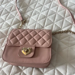 Rosa quiltad axelväska med hjärtspänne - Supersöt ljusrosa axelväska med quiltat mönster och guldfärgad kedja. Locket har ett hjärtformat spänne i guld som ger väskan en gullig touch. Liten och i perfekt storlek för bland annat en yngre tjej. Väskans storlek gemförs men ett mobilskal på sista bilden. Pris går att diskutera🥰