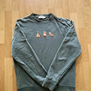 Grön sweatshirt från Maison Kitsuné - Säljer en grön sweatshirt från Maison Kitsuné i bomull med broderade rävar på bröstet. Tröjan har rund hals, långa ärmar och ribbade muddar. Perfekt för dig som gillar stilrena plagg med en twist. Storlek S.