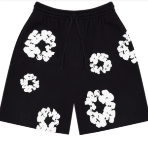 Svart denim tears  - Svarta shorts med elastisk midja och snörning. De har stora vita blommiga tryck över hela plagget som ger en cool och unik vibe. Passformen är loose och materialet känns som bomull, perfekt för varma dagar.