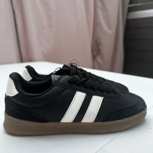 Adidas Samba svarta sneakers - Klassiska Adidas Samba sneakers i svart skinn med vita ränder och detaljer. Skorna har bruna gummisulor, svart snörning och rund tå. Perfekta för dig som gillar retrostil och streetwear. Helt ny