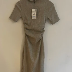 Beige långklänning med struktur - Snygg beige klänning med korta ärmar och diskret strukturerat tyg. Klänningen har en enkel, rak passform och lätt rynkade detaljer i midjan som ger en cool siluett. Perfekt för dig som gillar stilren och minimalistisk look.