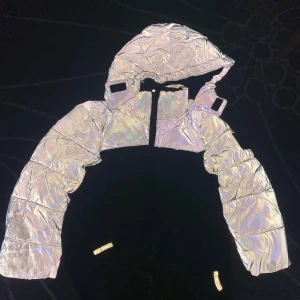 Reflekterande pufferjacka från WAVE - Säljer en riktigt cool pufferjacka från WAVE med reflekterande silverfärg på ärmar och huva. Jackan har dragkedja framtill och en schysst oversized look. Perfekt för dig som vill sticka ut och synas i mörkret.
