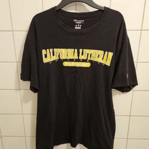 Svart Champion t-shirt California Lutheran - Vintage svart t-shirt från Champion med tryck i gult och vitt: 'California Lutheran University Grandparent'. Klassisk passform, korta ärmar och broderad Champion-logga på ärmen. Varan är äkta och köptes för 500 kr. Skriv gärna vid frågor och priset går såklart att diskutera.