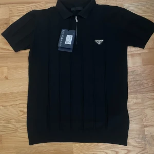 Svart pikétröja från Prada med dragkedja - Snygg svart pikétröja från Prada med klassisk krage, kort ärm och diskret logga på bröstet. Tröjan har en stilren dragkedja framtill och subtila vertikala ränder i tyget. Perfekt för dig som gillar exklusiv och stilren design.
