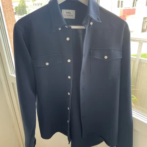 Mörkblå overshirt från Ciszere, slim S - Snygg mörkblå overshirt från Ciszere i slim fit. Skjortan har två bröstfickor med lock, vita knappar och en liten orange brodyr på ärmen. Materialet är en mix av polyester, viskos och elastan för en skön känsla och bra passform.