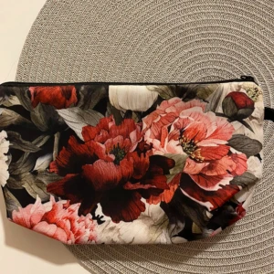 Blommig necessär med dragkedja - Necessär med stort blommönster i rött, rosa och vitt. Väskan har dragkedja upptill och är tillverkad i ett mjukt tygmaterial. Perfekt för smink eller småprylar när du är på språng.
