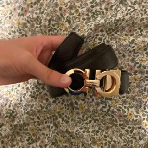 Svart Ferragamo skärp med gulddetalj - Snyggt svart skärp från Ferragamo i äkta skinn med ikoniskt guldspänne. Bältet har en stilren och klassisk design som lyfter vilken outfit som helst. Perfekt för dig som gillar exklusiva accessoarer och vill sticka ut med en lyxig detalj.