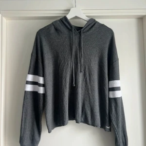 Grå hoodie från Hollister med ränder - Slutsåld vintage amerikansk baseball style grå ribbad hoodie från Hollister Must-Have Collection. Croppad modell med vita dubbla ränder på ärmarna, huva med snörning och lång ärm. Mjuk och skön, perfekt till chill dagar. Liten broderad logga nertill.