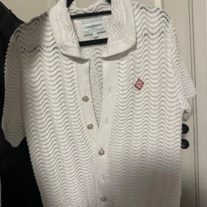 Casablanca Wavy crochet shirt - Säljer en vit stickad kofta från Casablanca med vågigt mönster, kort ärm och pärlknappar framtill. Koftan har en klassisk krage och röd broderad logga på bröstet. Perfekt för dig som gillar en clean och lyxig vibe.