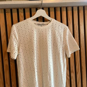 Vit broderad t-shirt från Zara - Vit t-shirt från Zara med korta ärmar och snyggt broderat hålmönster över hela plagget. T-shirten har rund halsringning och är gjord i mjuk bomull, perfekt för en fräsch och trendig look.