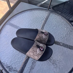 Sandaler med bi-motiv - Snygga sandaler med svart sula och bred rem i beige med klassiskt GG-mönster och coolt bi-tryck. Perfekta för sommaren och ger en lyxig vibe till din outfit. Remmen är i syntetmaterial och sulan är mjuk och bekväm.