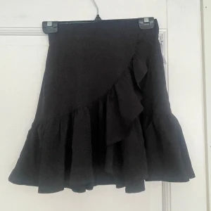 Svart volangkjol från Linne XS - Supersöt svart kortkjol från Linne i storlek XS. Kjolen har volanger och omlottkänsla framtill, vilket ger en flowy och trendig look. Perfekt att styla med en croppad topp eller oversized hoodie för en cool vibe.