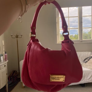 Röd axelväska från Marc by Marc Jacobs - Cool rosa axelväska i skinn från Marc by Marc Jacobs med guldiga metalldetaljer och ett brett handtag. Väskan har en innerficka med dragkedja. 