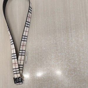 Burberry bälte  - Snyggt Burberry-bälte med klassiskt rutigt mönster i beige, svart, vitt och brunt. Bältet har ett stilrent metallspänne med TB-logga i silver. Tillverkat i skinn och passar perfekt för att lyfta din outfit med en lyxig touch.