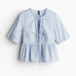 Ljusblå blus med knyt från H&M - Supersöt ljusblå blus från H&M med knyt framtill och volang nertill. Blusen har korta ärmar och är perfekt för vår och sommar. Den har en avslappnad passform.