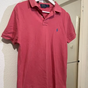 Rosa pikétröja från Polo Ralph Lauren - Klassisk rosa pikétröja från Polo Ralph Lauren med blå broderad logga på bröstet. Tröjan har korta ärmar, krage och knappar vid halsen. Perfekt för en stilren och avslappnad look.