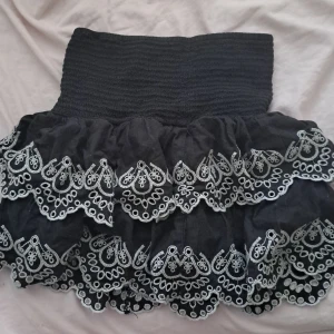 Svart volangkjol med broderad spets - Supersöt svart kortkjol med bred smock i midjan och flera lager volanger. Kjolen har vit broderad spets längs kanterna som ger en unik och lekfull look. Perfekt för dig som gillar att sticka ut med detaljer och volym.