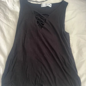 Svart ärmlös topp med snörning - Svart ärmlös topp från Nicole Simpson i mjuk bomull. Toppen har en snygg v-ringning med dekorativ snörning framtill som ger en cool touch. Perfekt att matcha med jeans eller kjol för en trendig look.