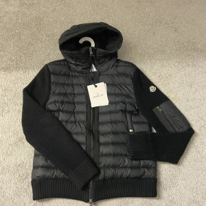Moncler Cardigan - Snygg svart pufferjacka från Moncler med stickade ärmar och quiltad framsida. Jackan har huva, dragkedja framtill och Moncler-logga på ärmen. Perfekt för dig som vill ha en stilren och sportig look med exklusiv känsla nu på sena sommarkvällar. Skick stabil 8.5/ 10 Nfc scan funkar. Hör av dig vid minsta lila tvekan så löser vi pris och andes frågor Mvh Emma 💗🤗