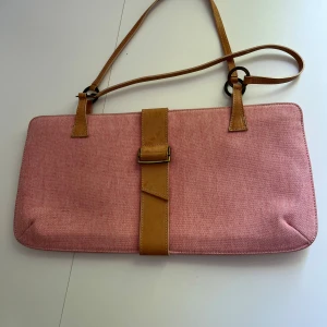 Rosa axelväska med skinnremmar - Säljer en snygg rosa axelväska i bomullstyg med ljusbruna detaljer och remmar i skinn. Väskan har en rektangulär form, justerbar axelrem och coolt spänne framtill. Perfekt för dig som vill ha en unik och stilren accessoar. Helt ny