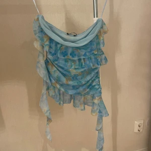 Blommig volang topp från Zara - Supersöt blommig topp från Zara i ljusblått med volanger och tunna axelband. Toppens meshmaterial ger en luftig känsla och de blå och gula blommorna gör den extra somrig. Perfekt för dig som gillar romantisk och trendig stil. Helt ny, liten i storlek.