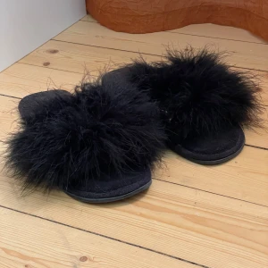 Svarta fluffiga tofflor med fjädrar - Svarta sandaler med bred rem täckt av mjuka, fluffig fjädrar. Platt sula med mjukistyg inne och gummisula. Passar bäst inomhus. Strl 39/40