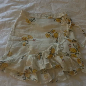 Blommig kjol med volang - Supersöt kort omlottkjol i vit med gula blommor och volangkant. Kjolen har knytband i sidan och är perfekt för en somrig vibe. Lätt och luftig känsla, passar perfekt till sneakers eller sandaler.