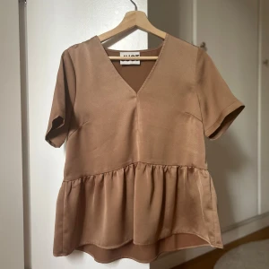 Beige blus med v-ringning från JUST - Supersöt beige blus från H&M med v-ringning och korta ärmar. Blusen har en volang nedtill som ger en flowy och avslappnad look. Perfekt till jeans eller kjol för en chill och trendig stil.