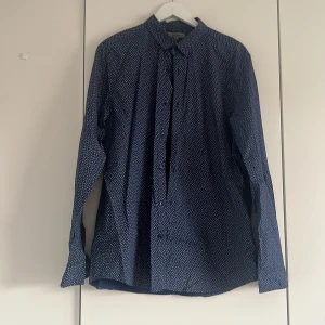 Blå blommig skjorta från S.C.W. - Snygg blå skjorta från S.C.W. i storlek XL med små vita blommor över hela tyget. Skjortan har klassisk krage, långa ärmar och knäppning framtill. Perfekt för dig som vill ha en stilren men ändå unik look.