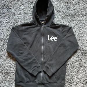 Svart hoodie från Lee med dragkedja - Svart hoodie från Lee med vit logga på bröstet. Tröjan har huva, hel dragkedja framtill och två fickor. Tillverkad i mjuk bomullsmix, perfekt för en chill och avslappnad stil. Regular fit och långa ärmar.