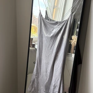 Glittrig silver klänning med tunna band - Supersnygg klänning i glittrigt silver med tunna axelband som korsas i ryggen. Klänningen har en draperad urringning och ett mjukt, följsamt tyg. Bandet är något trasigt men går lätt att sy på själv.