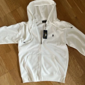Vit hoodie från Polo Ralph Lauren M - Snygg vit hoodie från Polo Ralph Lauren med dragkedja och huva. Klassisk svart broderad logga på bröstet och två fickor framtill. Tillverkad i mjuk bomull, perfekt för en clean och stilren look.