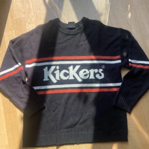 Svart stickad tröja från Kickers - Svart stickad tröja från Kickers med vita och röda ränder över bröstet och ärmarna. Stor logga framtill. Klassisk rund halsringning och ribbade muddar vid ärmslut och nederkant. Perfekt för dig som gillar retrovibbar och streetstyle.