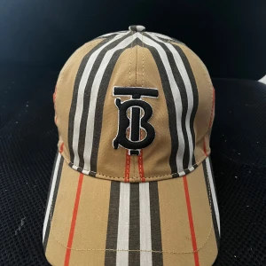 Burberry keps - Snygg keps från Burberry med klassiskt randigt mönster i beige, svart, vitt och röda detaljer. Framtill finns en broderad TB-logga i svart och vitt. Kepsen är tillverkad i 100% bomull och har justerbar rem baktill för perfekt passform.