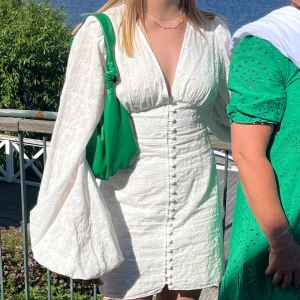 Vit kortklänning från Adoore - Superfin vit kortklänning från Adoore med broderade blommor, djup v-ringning och långa puffärmar. Klänningen har knappar framtill och markerad midja för en snygg siluett. Perfekt för sommarens alla tillfällen!