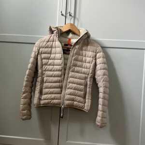 Beige dunjacka från Parajumpers YS - Snygg beige dunjacka från Parajumpers i storlek YS. Jackan har huva, två dragkedjefickor och klassisk PJS-logga på ärmen. Lättviktig och quiltad design som ger en clean och modern look. Perfekt för kyliga dagar när du vill hålla stilen. Använd och har defekter (hål, 2st) Som är lappade (rygg, ficka)