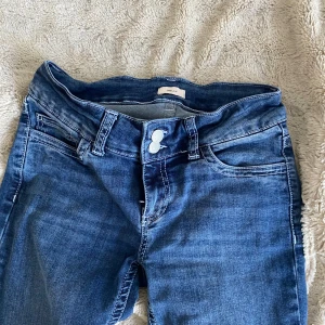 Blå bootcut jeans från Nelly - Snygga blå bootcut jeans från Nelly med klassisk femficksdesign och dubbla knappar i midjan. Jeansen har dekorativa vita sömmar på bakfickorna och är tillverkade i mjukt denimtyg. Perfekt för dig som gillar en retroinspirerad look. Säljer då dom aldrig har kommit till användning 