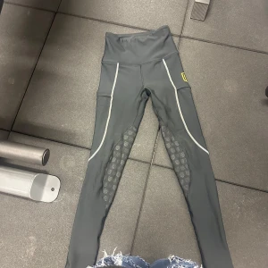 Svarta ridleggings från Maya Delorez - Snygga svarta ridleggings från Maya Delorez med hög midja och gråa detaljer längs benen. Silikonprint på insidan av benen för extra grepp och gult logomärke på låret. Tillverkade i stretchigt syntetmaterial som andas och sitter tajt. Väldigt bra kvalité på byxorna och har ej andvändt mycket, säljer pga att jag slutat i ridning. Nypris: 899kr