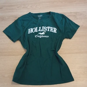 Grön Hollister t-shirt med tryck - Mörkgrön t-shirt från Hollister med vit logga och texten 'California' på bröstet. Klassisk rund hals och korta ärmar. Tillverkad i 100% bomull för en skön och avslappnad känsla. Perfekt för dig som gillar enkel och stilren streetwear.