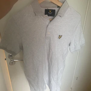 Ljusgrå pikétröja från Lyle & Scott - Snygg ljusgrå pikétröja från Lyle & Scott med klassisk krage och korta ärmar. Tröjan har den ikoniska gula örnloggan broderad på bröstet och knäppning framtill. Perfekt för en clean och stilren look.