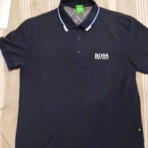 Marinblå pikétröja från Hugo Boss - Snygg marinblå pikétröja från Hugo Boss med vit logga på bröstet och ryggen. Klassisk krage med vita och blå ränder, knäppning framtill och korta ärmar. Perfekt för en clean och stilren look.