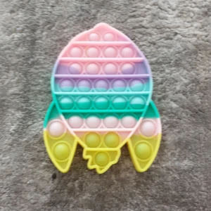 En raket poppit i massa färger - Bilderna visar en färgglad pop it-leksak i form av en raket, inte en bok. Den har pastellfärger i rosa, lila, grön och gul. Produkten är avsedd för stresslindring och lek, men är inte relevant för bokkategorier.