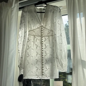 Vit blommig kortklänning från Adoore - Superfin vit kortklänning från Adoore med broderade blommor, v-ringning och långa puffärmar. Klänningen har knappar framtill och markerad midja för en snygg siluett. Perfekt för sommarens alla tillfällen!