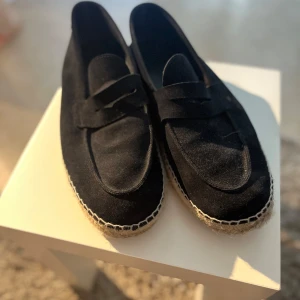 Svarta mockaloafers med flätad sula - Snygga svarta loafers från Massimo i mocka med klassisk pennyloafers-design. Skorna har en rund tå och en flätad sula i jute för en chill sommarkänsla. Perfekta att glida runt i när det är varmt ute. Behöver tilläggas det krävs att man både har långa och bredda fötter för dessa.