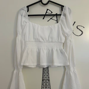 Vit blus med volang och puffärm från Gina Tricot - Superfin vit blus med volangkant och smockad midja från Gina Tricot i storlek XS. Toppen har långa puffärmar med resår och en fyrkantig ringning. Tunt, strukturerat tyg som ger en luftig känsla. Aldrig använd. 