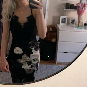 Svart blommig spetsklänning med smala axelband - Säljer en svart klänning med smala axelband, spets upptill och stora blommor nertill. Klänningen har v-ringning och fina spetsdetaljer över bysten. Kjoldelen har ett stort blommönster i vitt och beige. Perfekt för sommarens tillfällen!