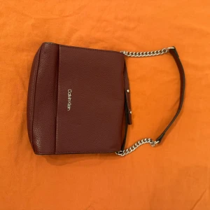 Vinröd axelväska från Calvin Klein - Snygg vinröd axelväska från Calvin Klein i strukturerat skinn. Väskan har en stilren design med silverfärgad kedja och logga framtill. Perfekt storlek för det viktigaste och ger en clean look till din outfit.