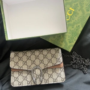 Dionysus axelväska med kedja - Väskan är beige och brun med bruna detaljer och insida i ljust skinn. Perfekt storlek. Helt oanvänd. 