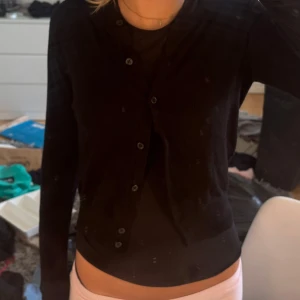 Svart kofta med knappar - En klassisk svart kofta med långa ärmar och knäppning framtill. Enkel och stilren design som passar till det mesta. Koftan har ribbade muddar vid ärmsluten och är i ett mjukt material som känns skönt mot huden.